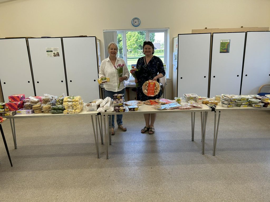 two women standing behind a table covered in food from marks and spencers including sweetcorn, bread, salad.