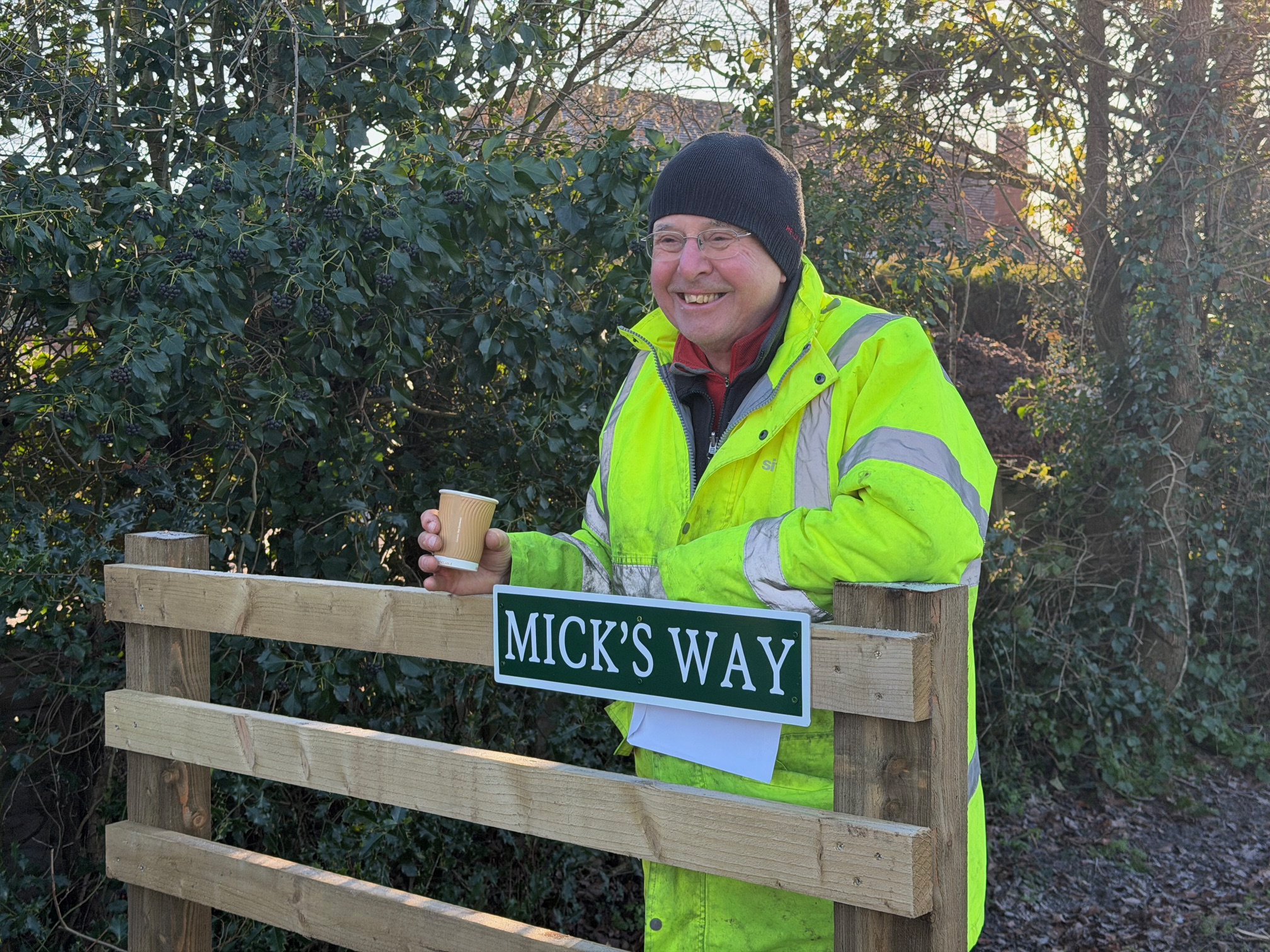 Photo of a gentleman (Mick Davies) wearing high vis jacket standing next to a footpath sign labelled Mick's Way