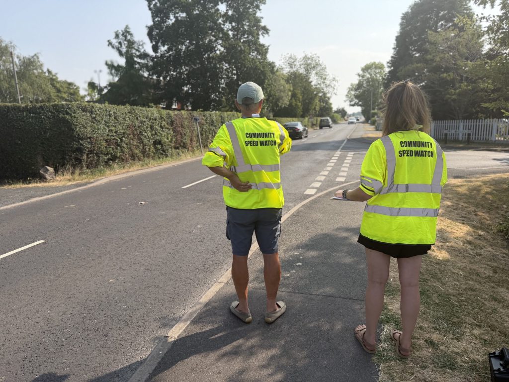 two people in high vis jackets with the words community speedwatch standing by the side of a road - you. can see their backs as they monitor traffic speeds
